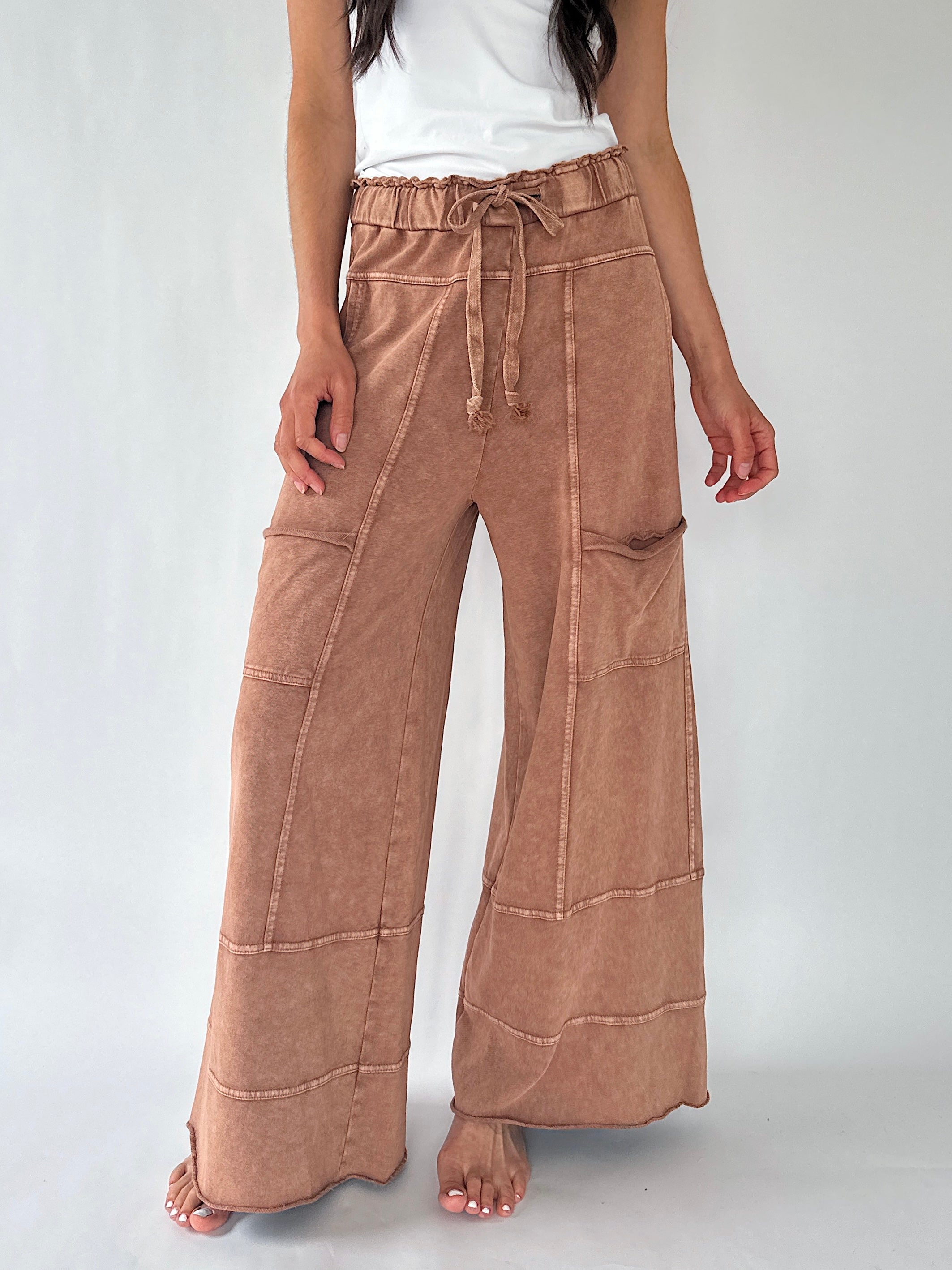A model wearing a pair of wide-leg, high-waisted pants in a dusty tan or light brown color, styled with a white sleeveless top against a light gray studio background.