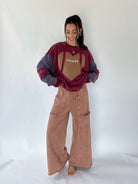 A woman modeling a football-themed sweatshirt and brown wide-leg pants against a plain white studio backdrop. The sweatshirt is burgundy and gray with a tan football patch on the chest, and the pants are a dusty rose-brown color.