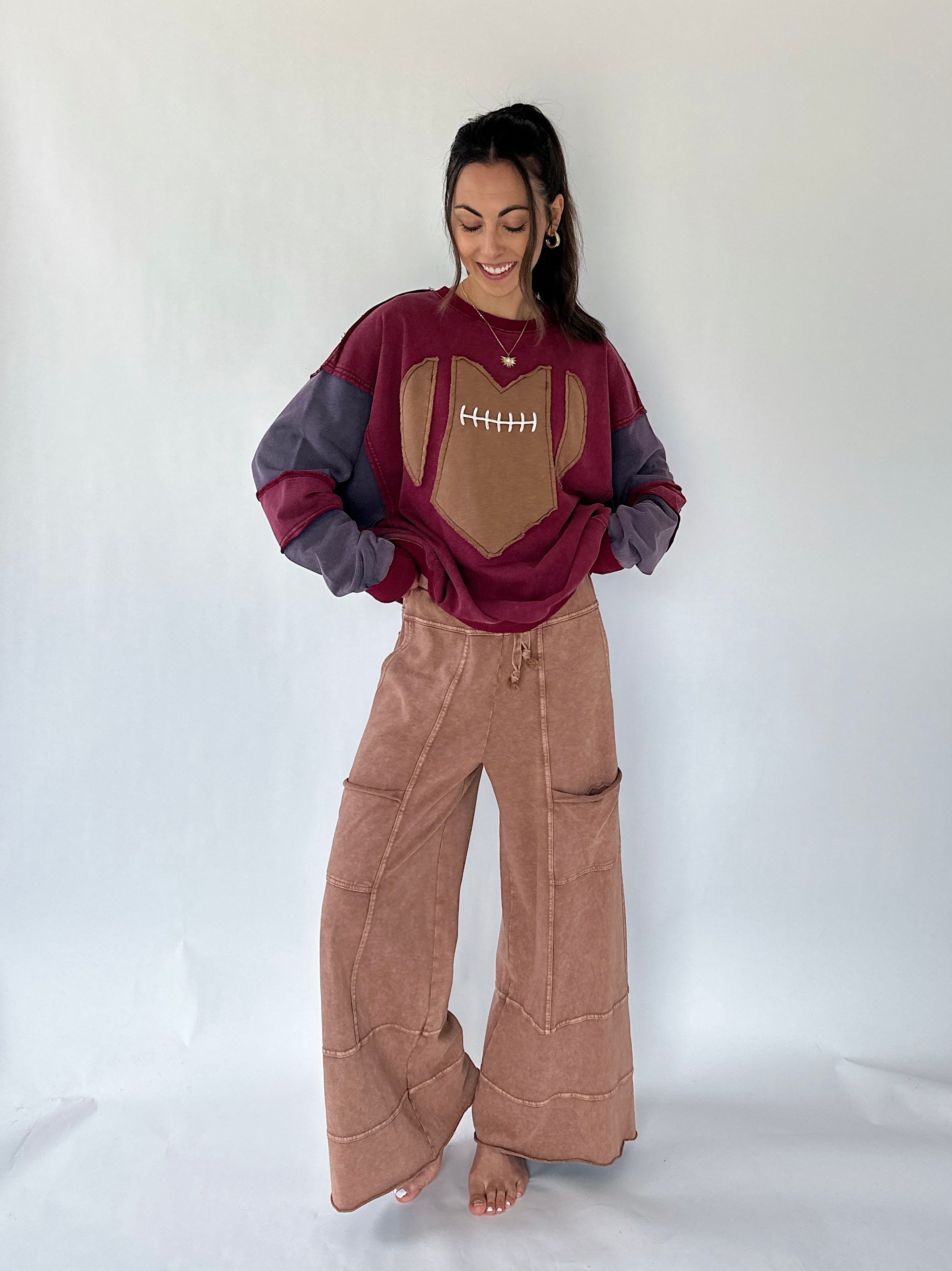 A woman modeling a football-themed sweatshirt and brown wide-leg pants against a plain white studio backdrop. The sweatshirt is burgundy and gray with a tan football patch on the chest, and the pants are a dusty rose-brown color.