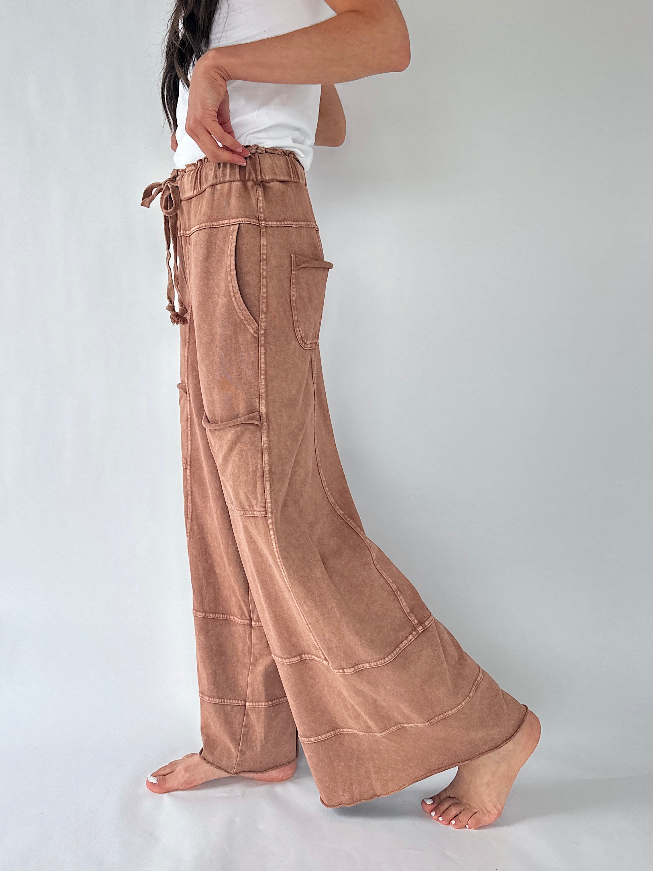A person wearing loose, wide-leg brown pants with a drawstring waist and large patch pockets, standing barefoot against a plain light gray studio background.