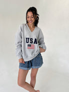 Woman wearing a gray sweatshirt with 'USA' and an American flag design, standing against a plain background.