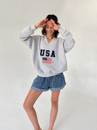 Woman wearing a gray sweatshirt with 'USA' and an American flag design, standing against a plain background.