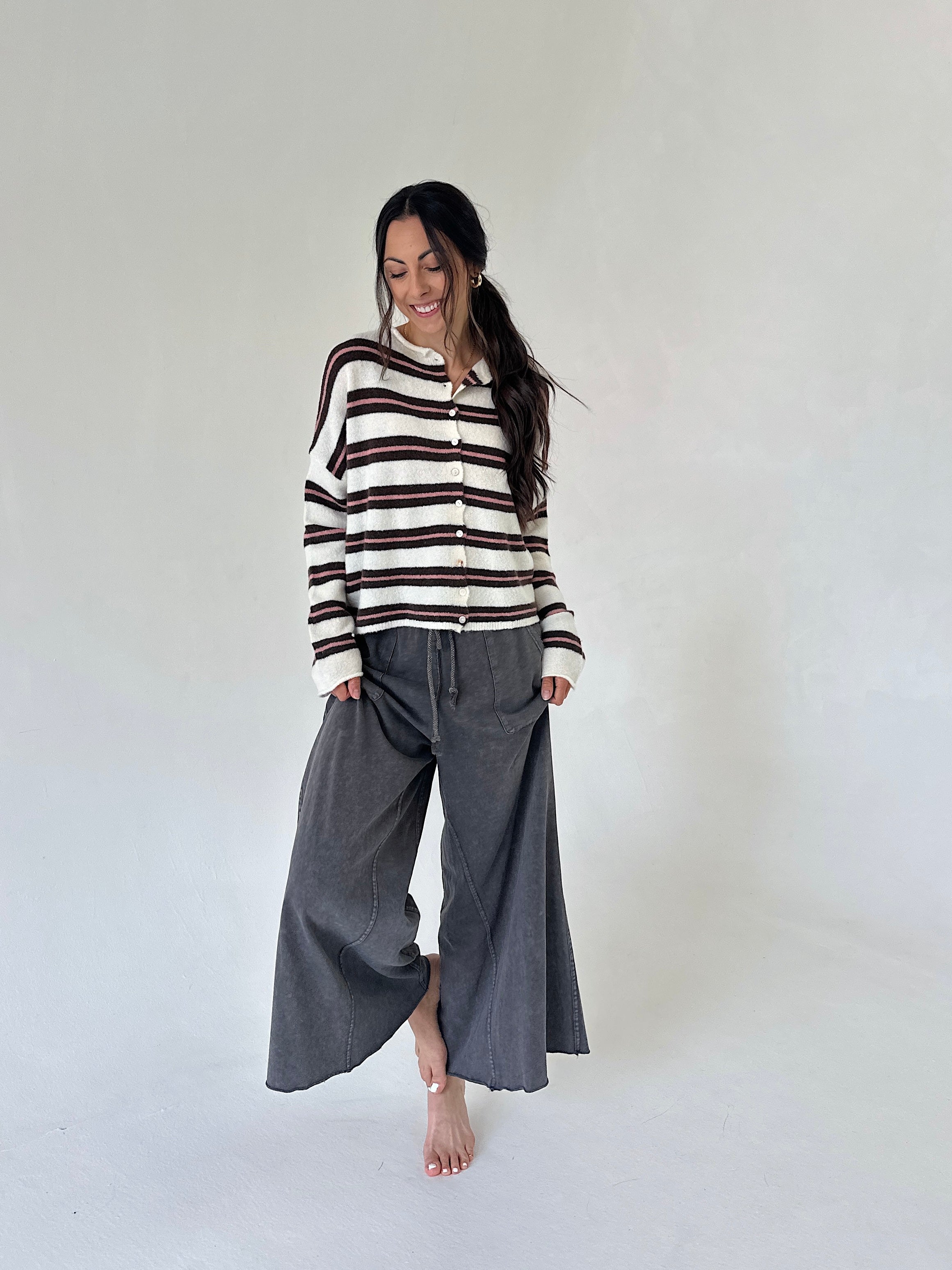 A woman stands barefoot against a plain light gray studio background, wearing a cream knit cardigan with dark brown horizontal stripes and loose dark gray wide-leg pants.