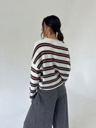 A woman is shown from behind wearing a cream-colored knit sweater with dark brown and pink horizontal stripes, paired with wide-leg gray pants, against a plain off-white studio background.