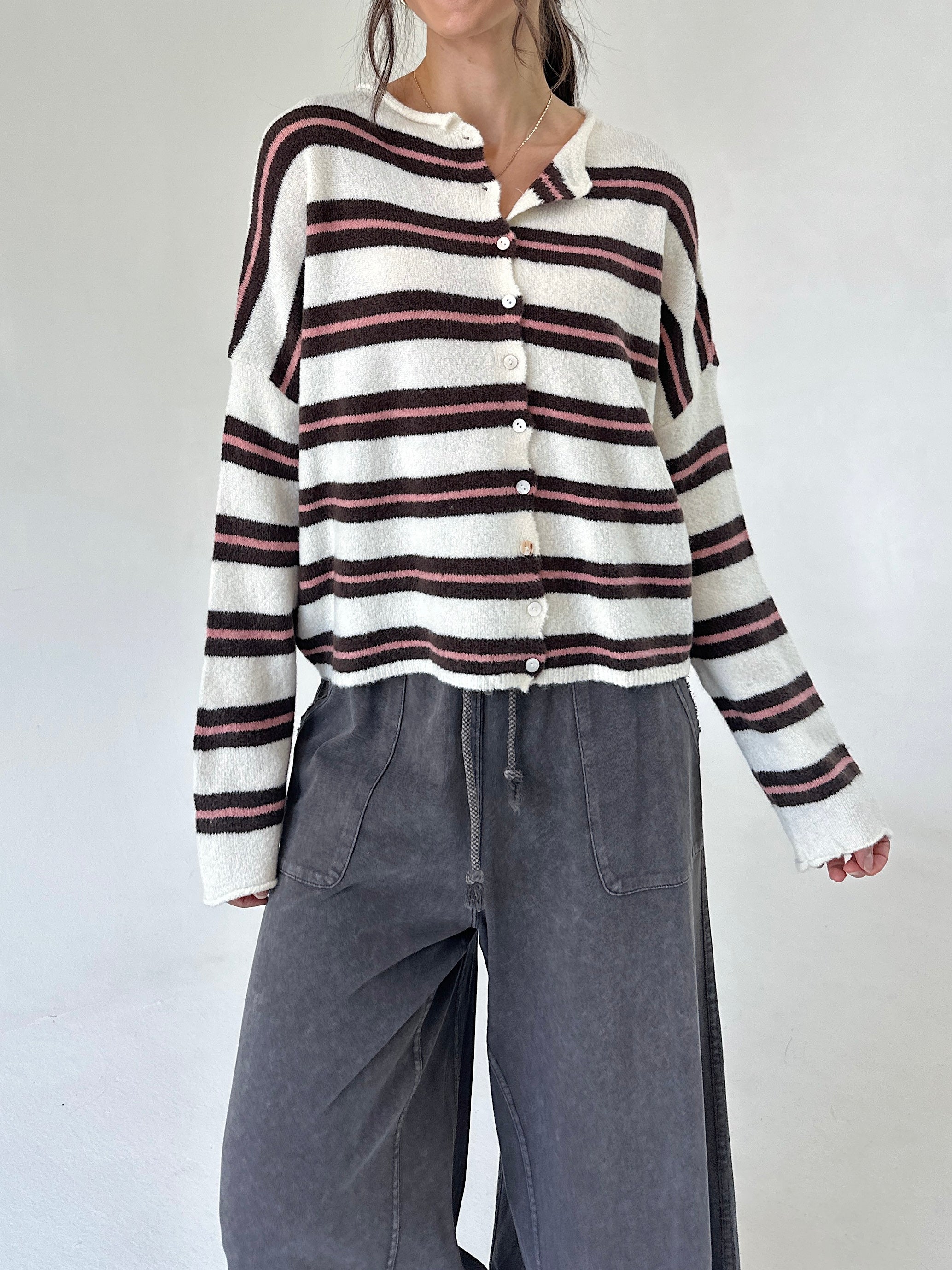 A woman wearing a cream knit button-up cardigan with dark brown and pink horizontal stripes, paired with wide-leg charcoal gray pants, against a plain light gray background.