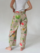 A person shown from the back wearing beige wide-leg pants with a colorful vegetable print, paired with a white t-shirt, against a light gray studio background.
