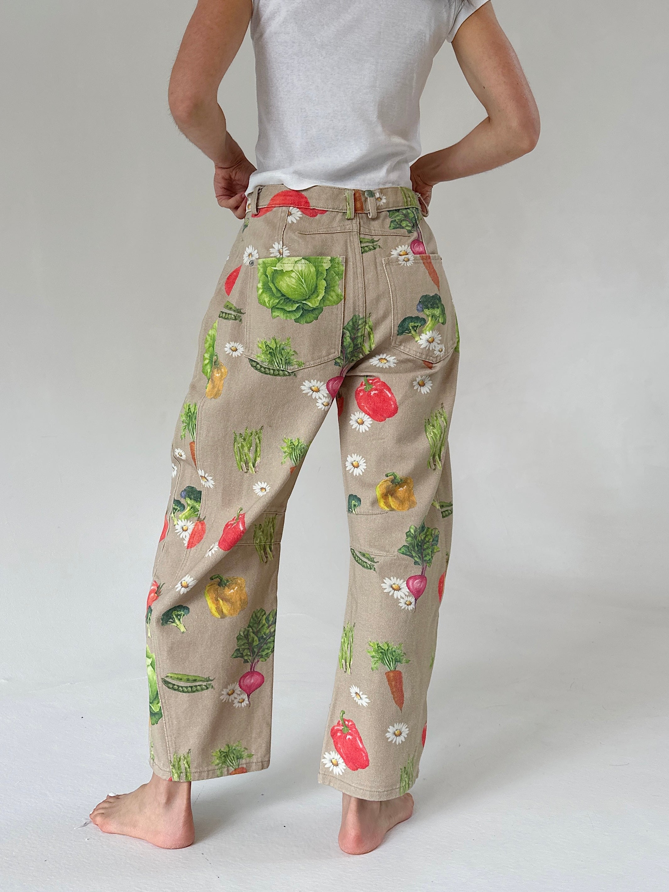 A person shown from the back wearing beige wide-leg pants with a colorful vegetable print, paired with a white t-shirt, against a light gray studio background.
