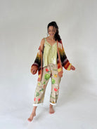 A woman modeling a light green printed tank top and beige cropped pants with a colorful vegetable print, styled with a multicolor striped knit cardigan, against a plain light gray studio background.