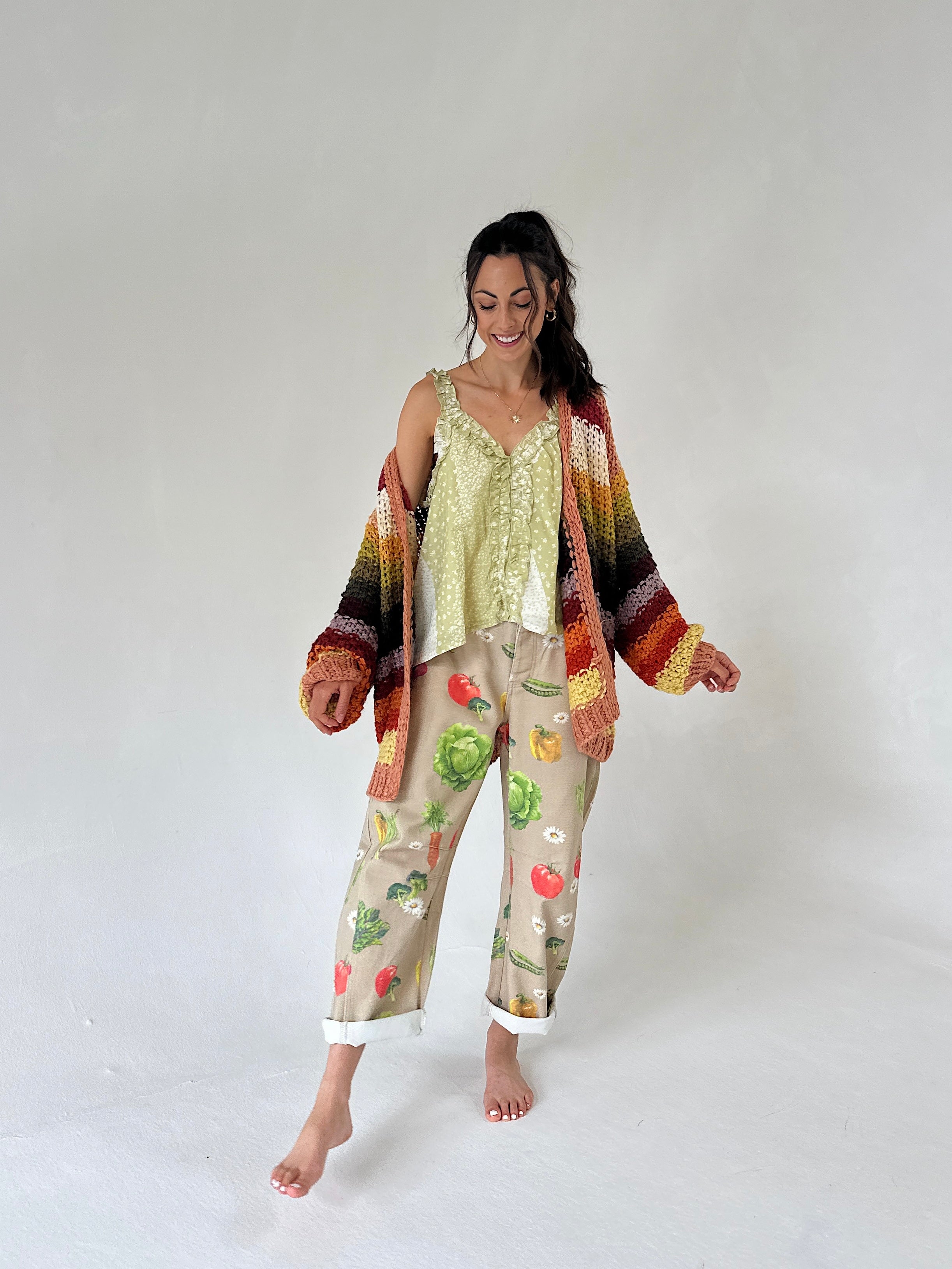 A woman modeling a light green printed tank top and beige cropped pants with a colorful vegetable print, styled with a multicolor striped knit cardigan, against a plain light gray studio background.
