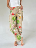 A person wearing beige wide-leg pants with a colorful vegetable print, legs crossed, paired with a white tank top, shown against a light gray studio background.