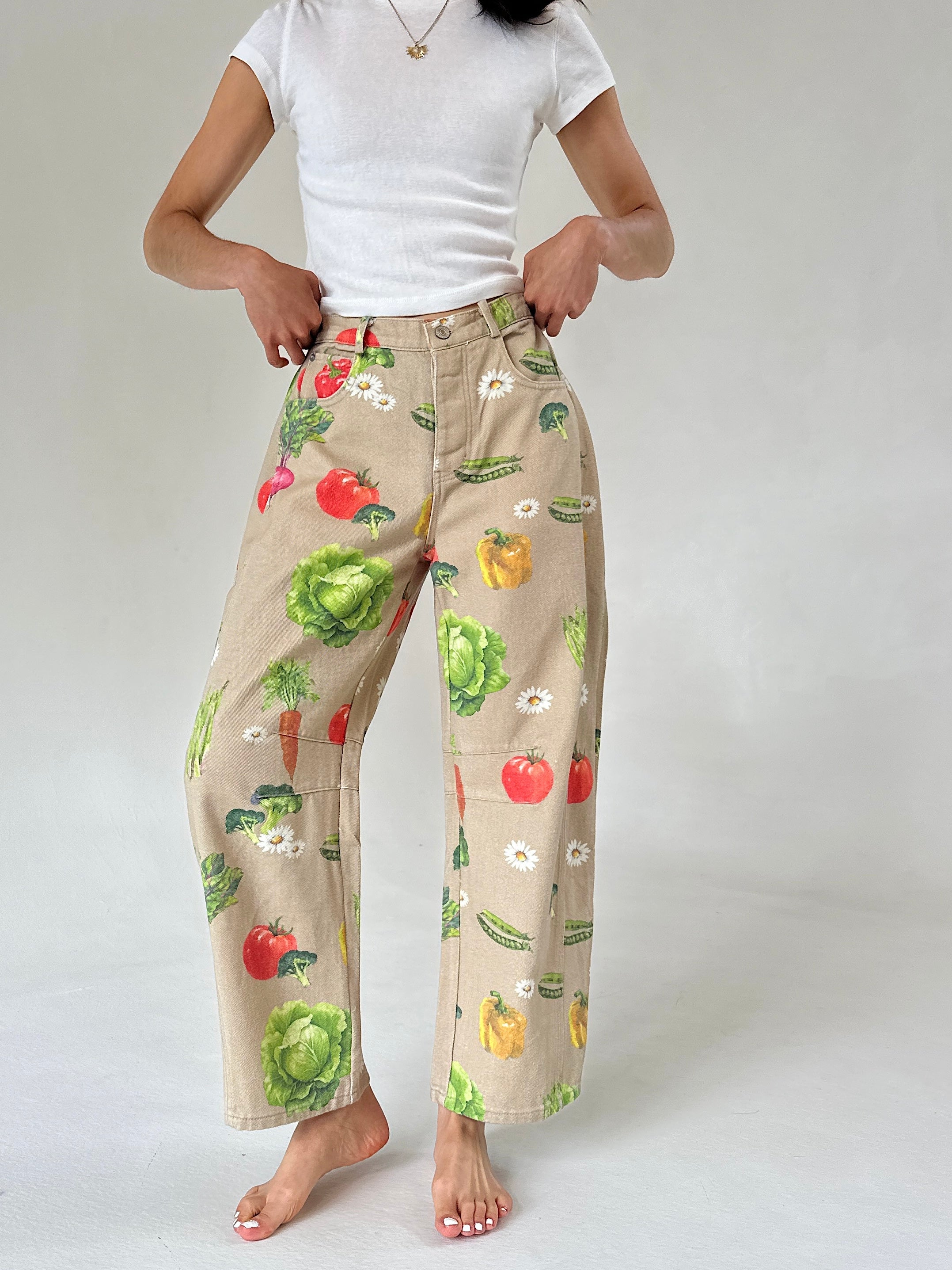 A person wearing beige wide-leg barrel pants with a colorful vegetable print, paired with a white t-shirt, standing against a light gray studio background.