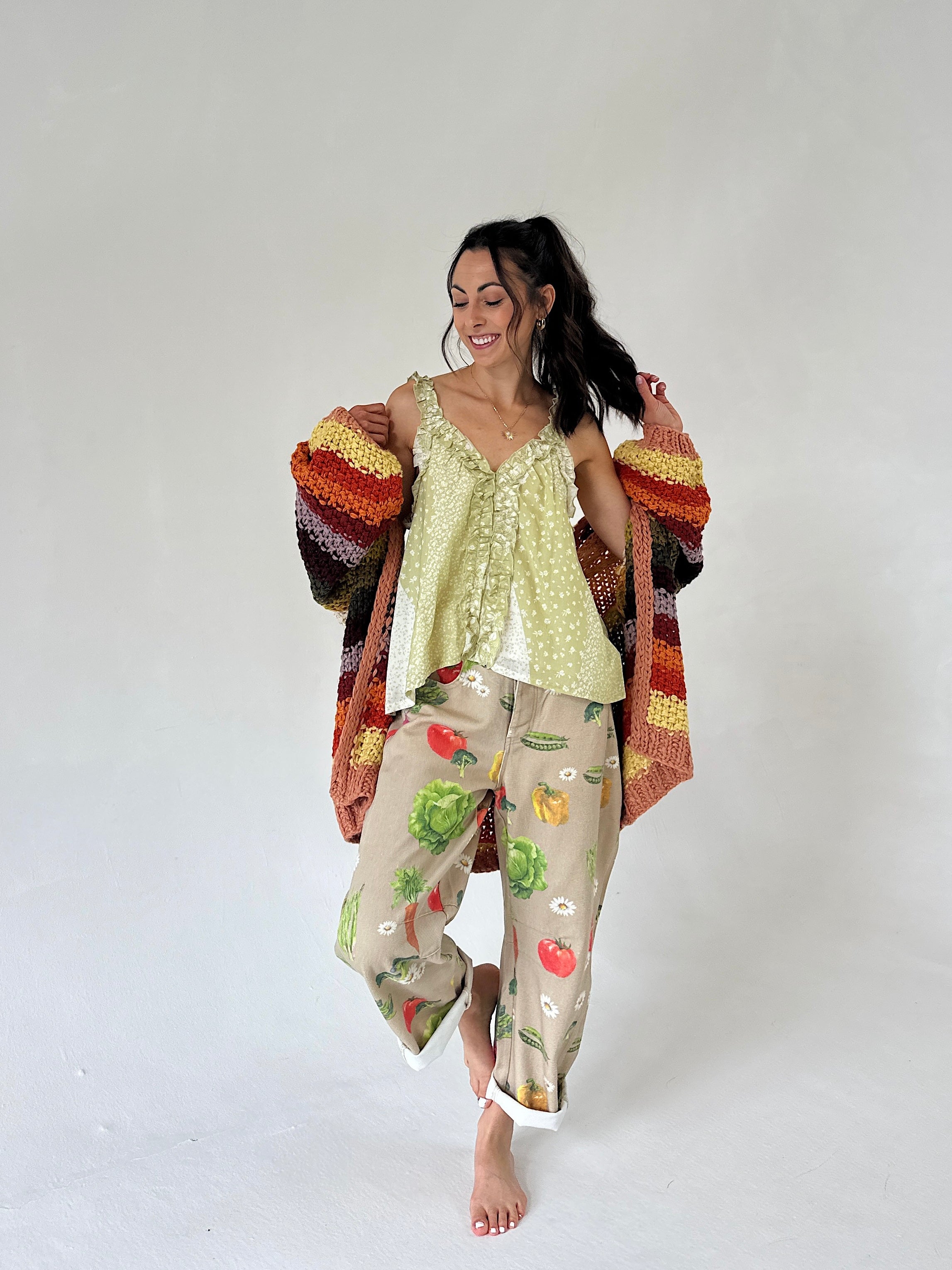 A woman modeling a light green ruffled tank and beige pants featuring a colorful vegetable print, styled with a striped knit cardigan against a plain light gray background.