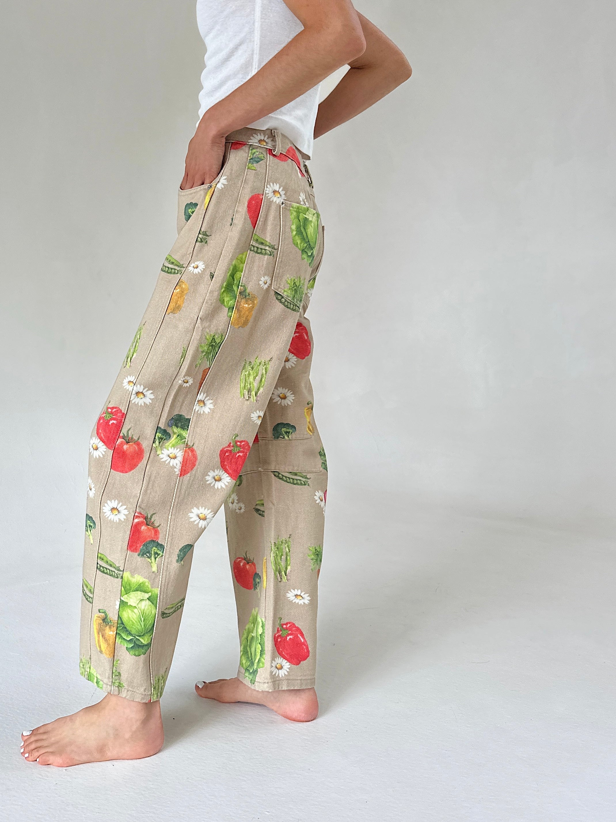 A person wearing beige wide-leg pants with a colorful vegetable print, standing barefoot against a light gray studio background.