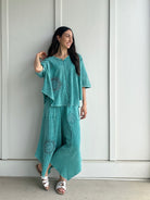 Woman wearing a teal outfit with patterns against a white paneled wall.