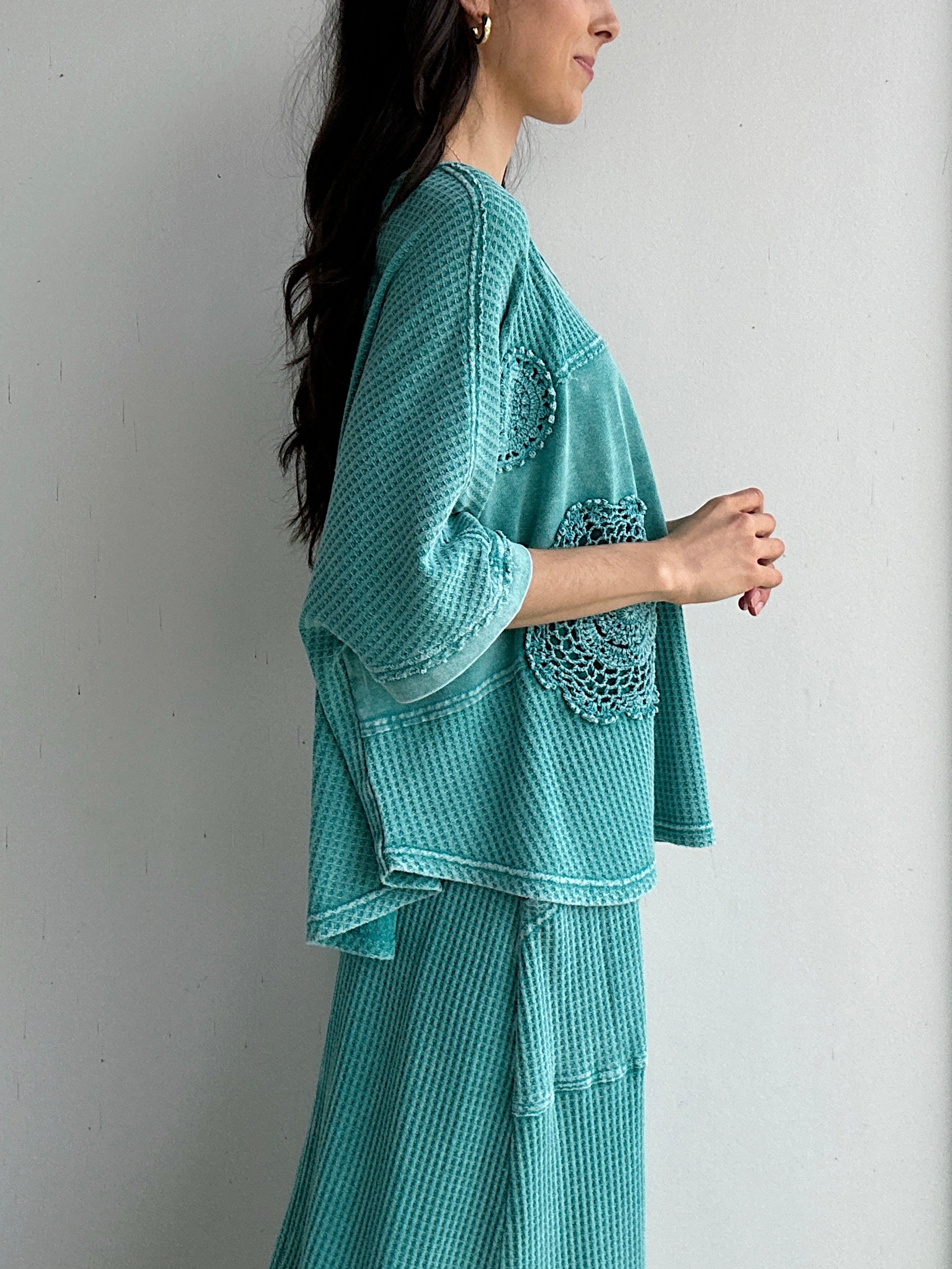 Woman wearing a teal textured outfit with lace details against a plain background showing side