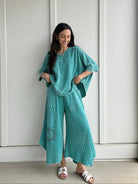Woman wearing a teal outfit with a matching top and wide-leg pants against a white wall.