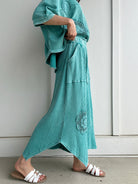 Person wearing a teal outfit with embroidered details against a white wall showing side