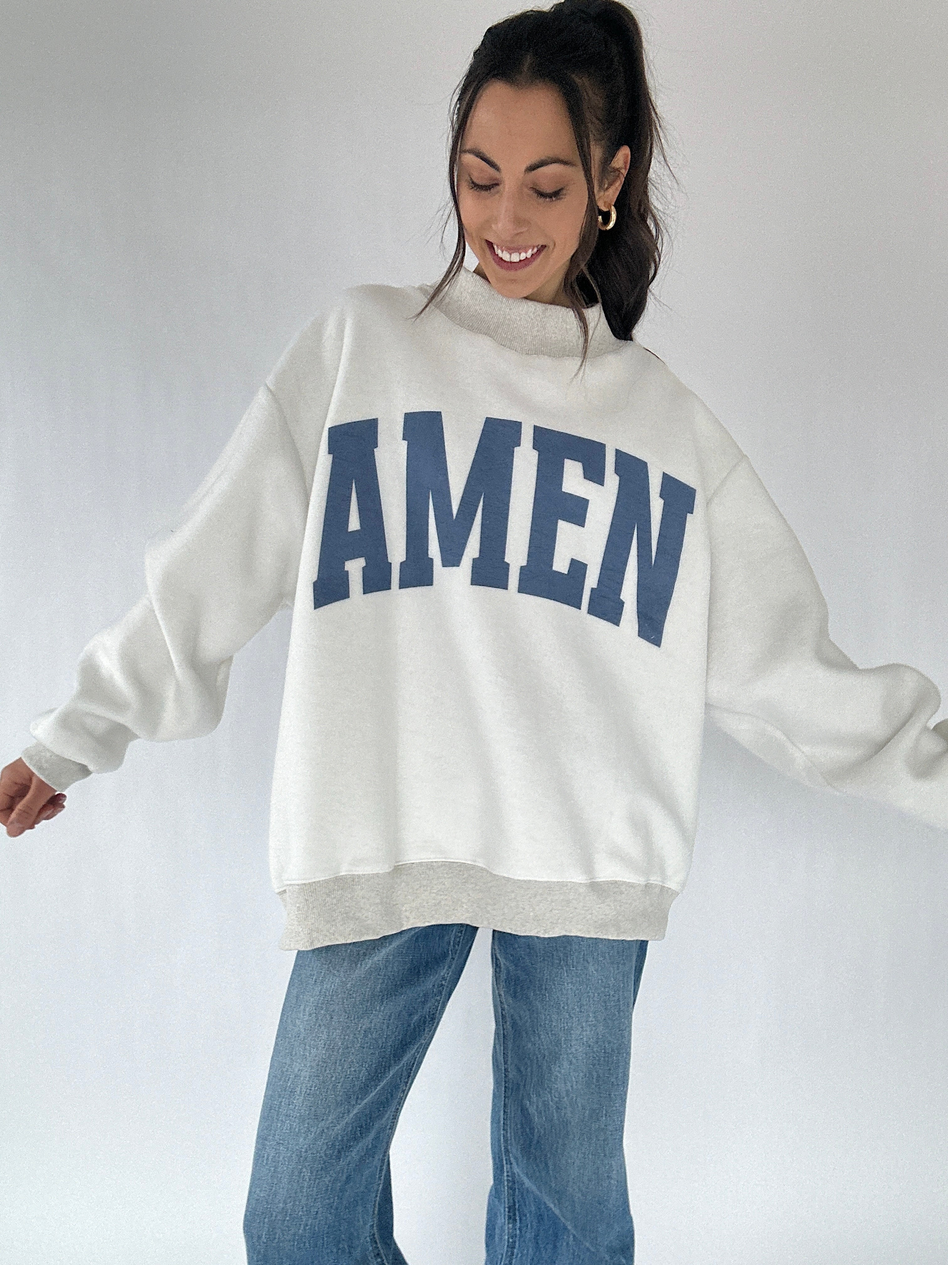 Woman wearing a white sweatshirt with 'AMEN' printed in blue on a plain background