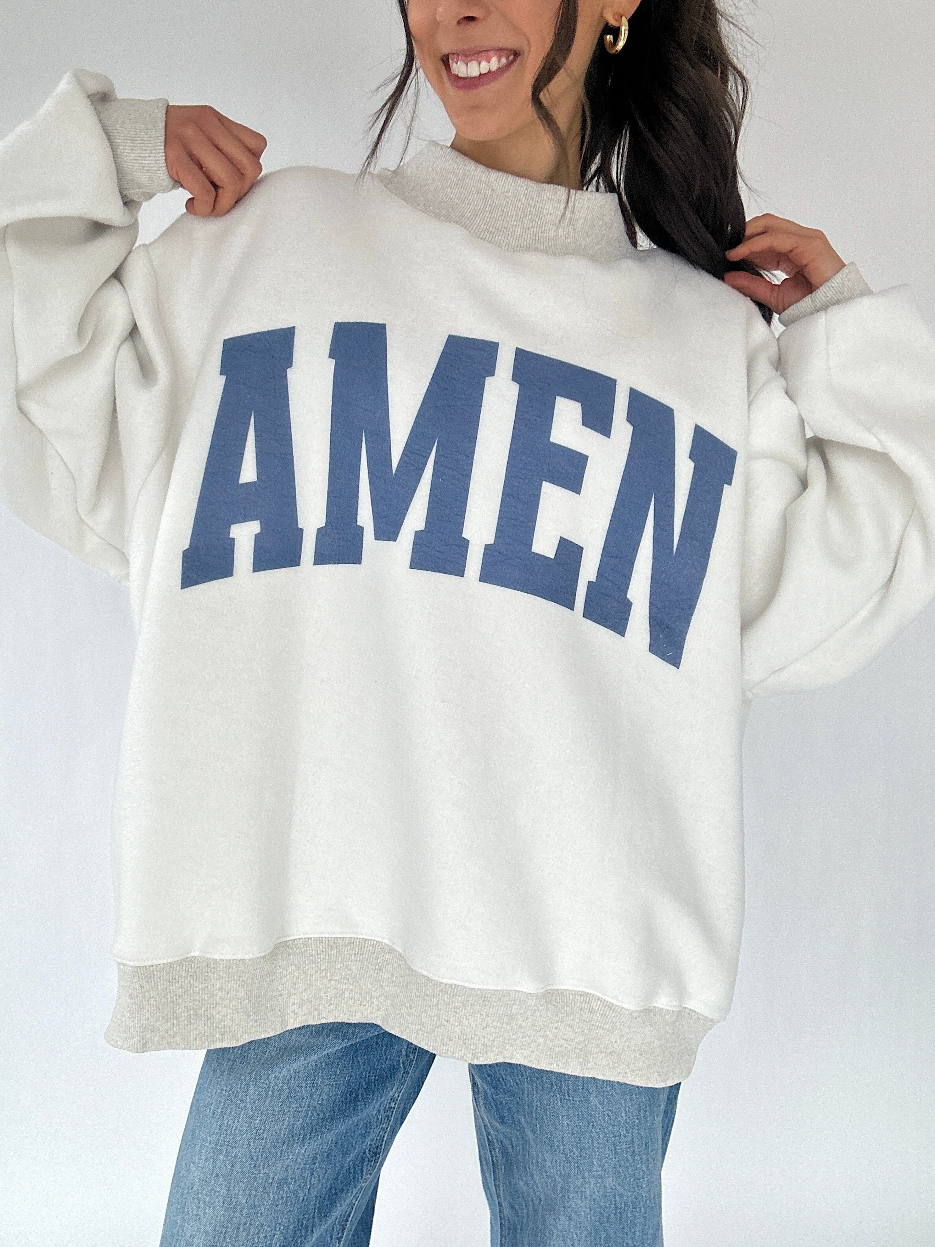 Person wearing a white sweatshirt with 'AMEN' printed in blue on a plain background