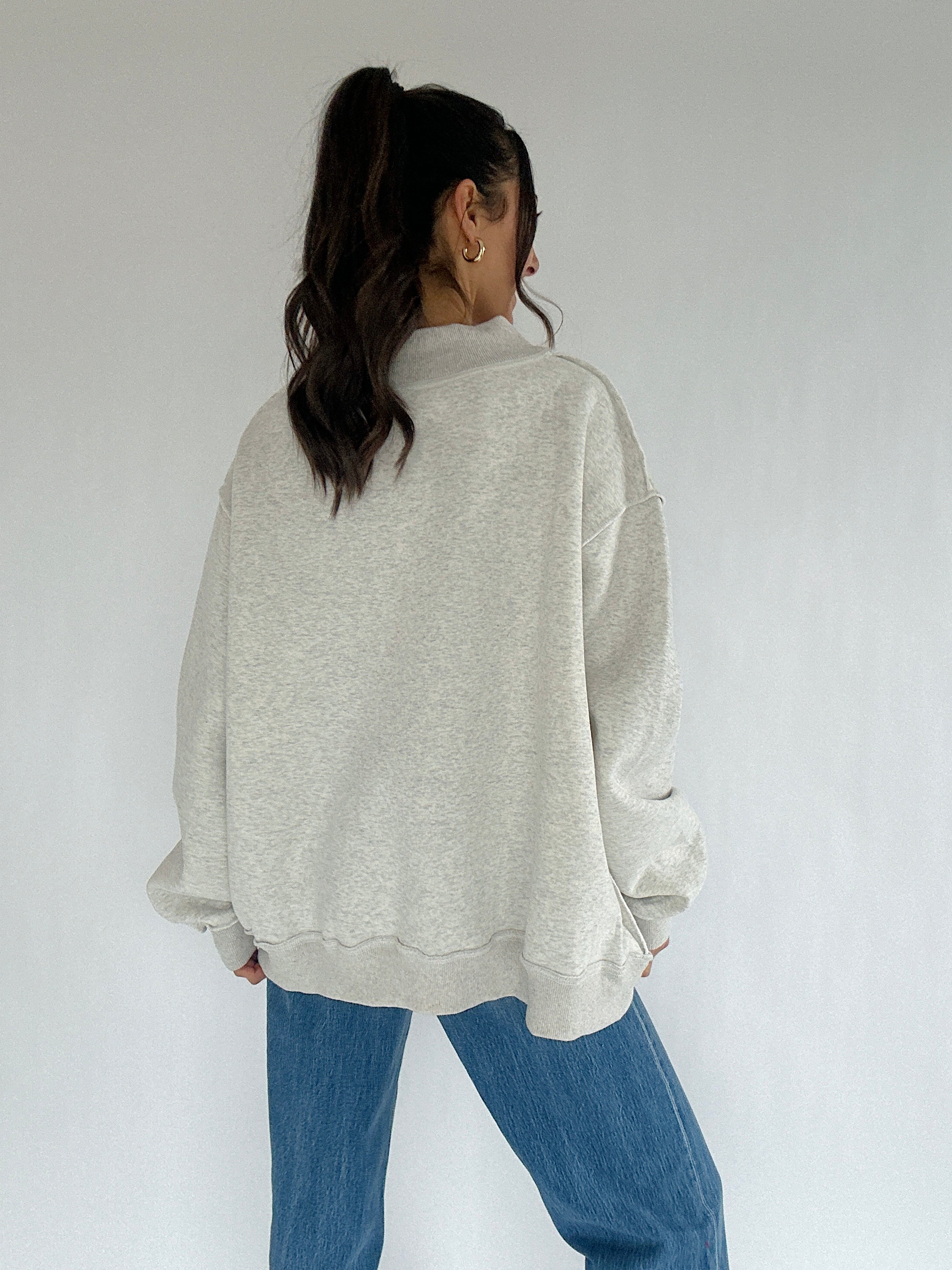 Person wearing a light gray sweatshirt and blue jeans against a white background showing back