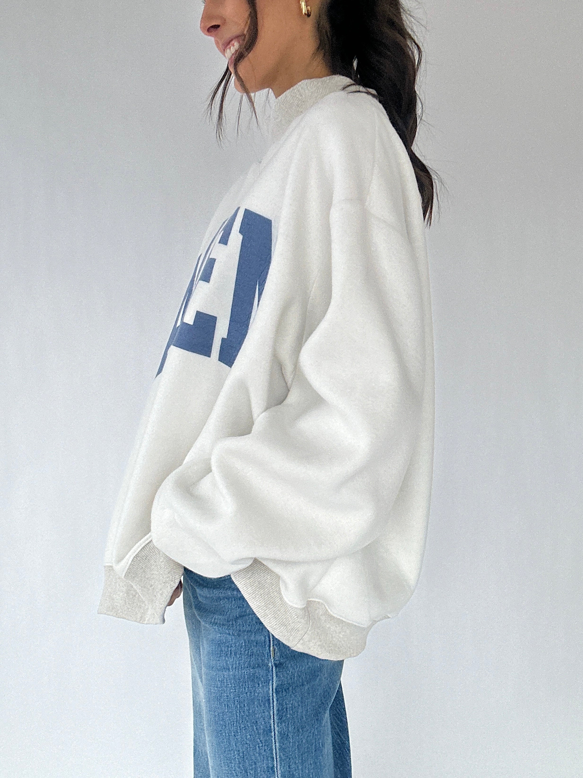 Person wearing a white sweatshirt with blue text and blue jeans on a plain background showing the side