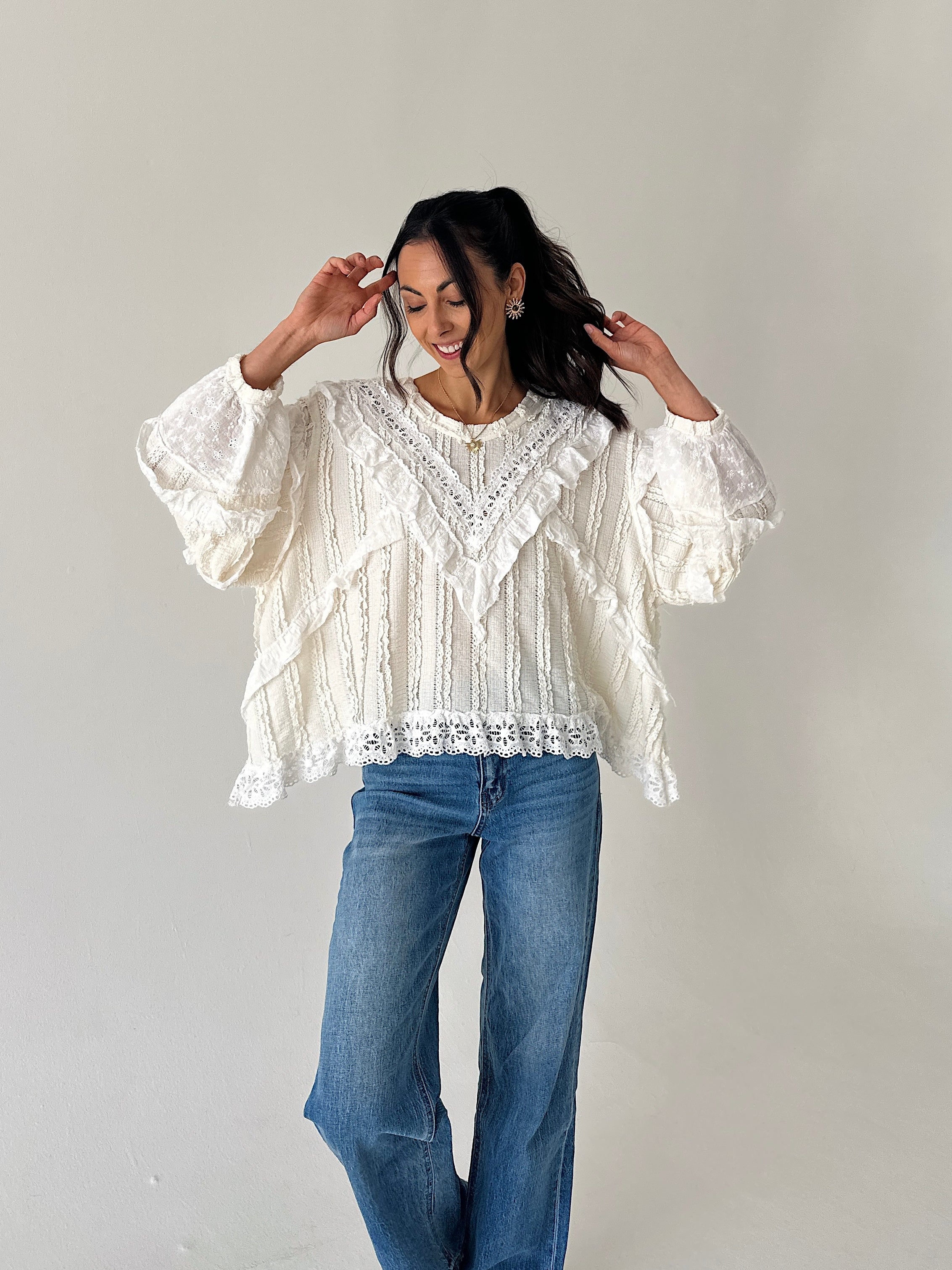 A woman modeling a white lace blouse with ruffles and blue jeans, standing against a plain light gray studio background.
