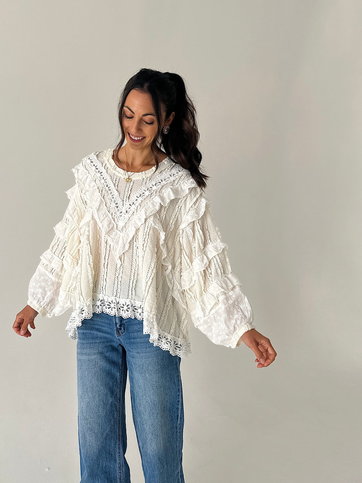 A woman wearing a white ruffled lace blouse with blue jeans, standing against a plain light gray studio background.