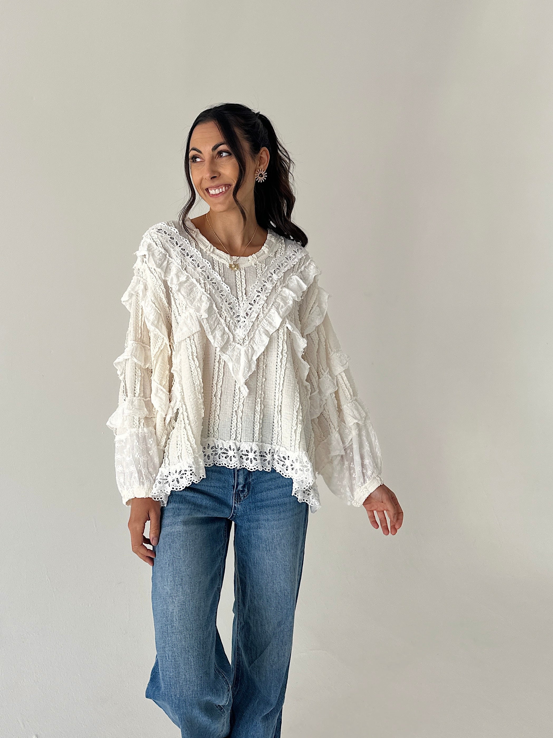 A woman modeling a cream-colored, long-sleeved bohemian-style blouse with intricate lace and ruffle details, paired with blue jeans, against a plain light gray background.