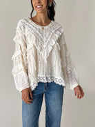 A woman wearing a cream-colored, long-sleeved lace blouse with ruffles and blue jeans, standing against a plain light gray wall.