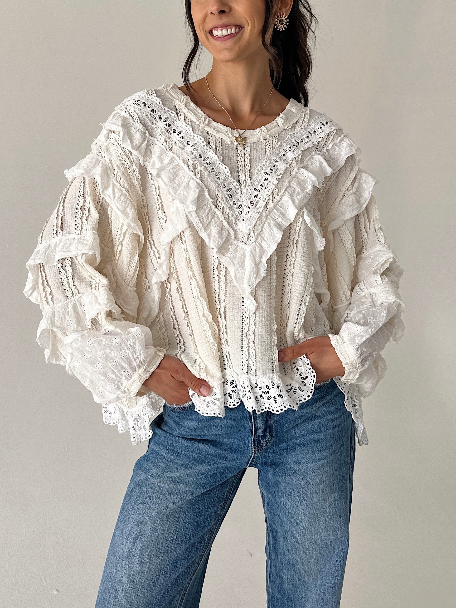A woman wearing a cream-colored, heavily ruffled and lace-trimmed long-sleeve blouse paired with blue jeans, standing against a plain light gray wall.