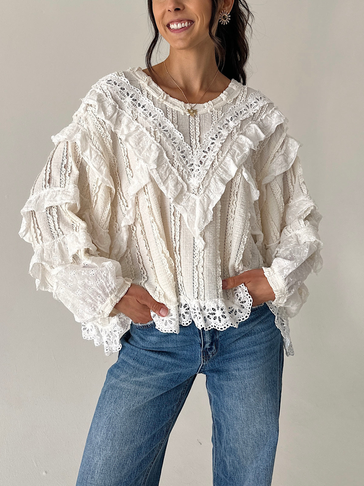 A woman wearing a cream-colored, heavily ruffled and lace-trimmed long-sleeve blouse paired with blue jeans, standing against a plain light gray wall.