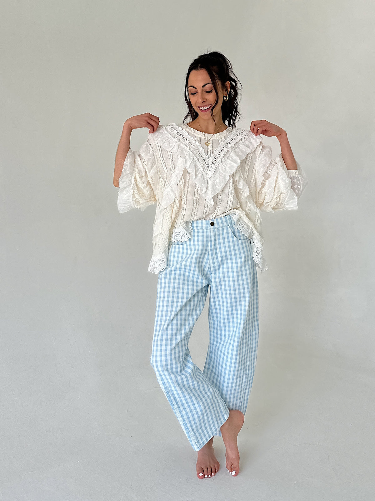 Woman wearing a white lace top and light blue checkered pants against a plain background