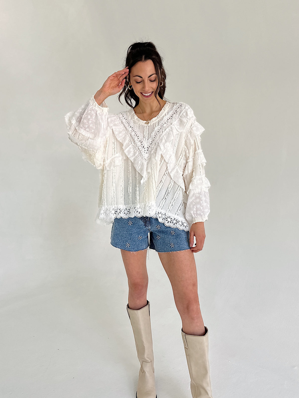 Woman wearing a white lace blouse, denim shorts, and beige knee-high boots against a plain background