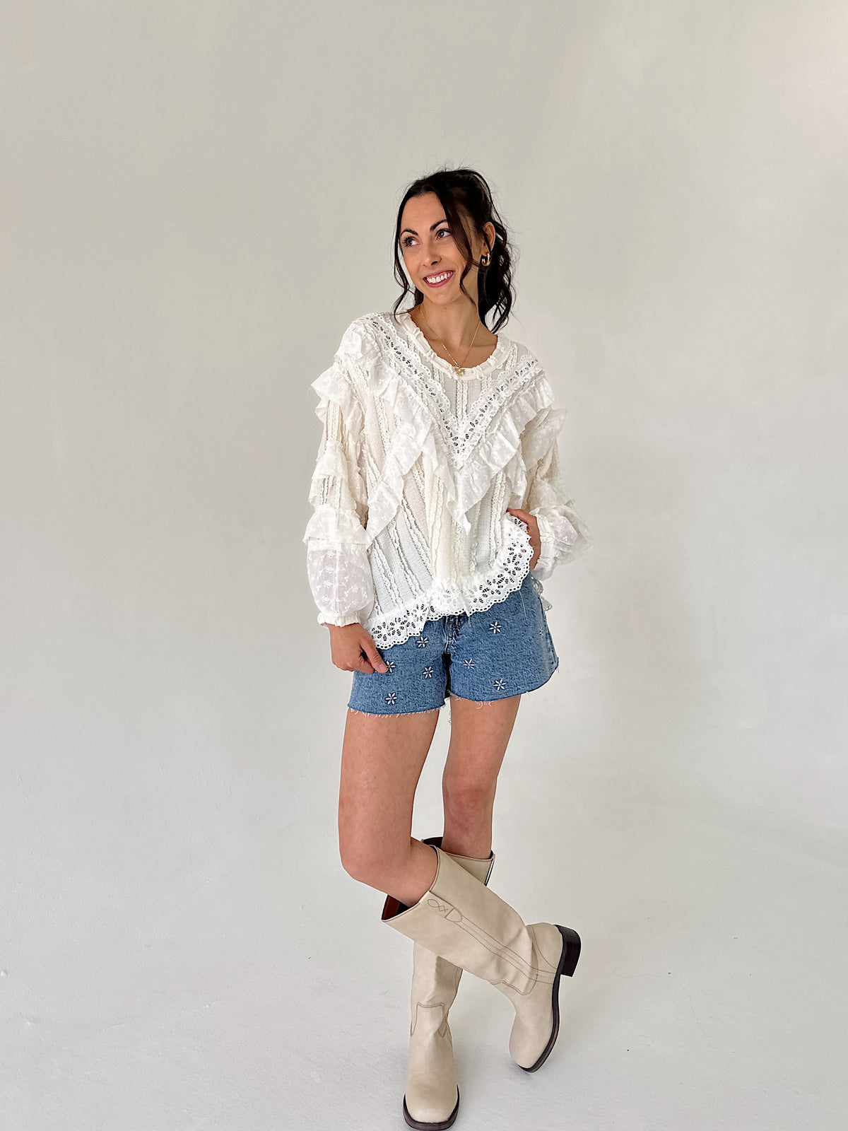 Woman wearing a white lace blouse, denim shorts, and beige knee-high boots against a plain background
