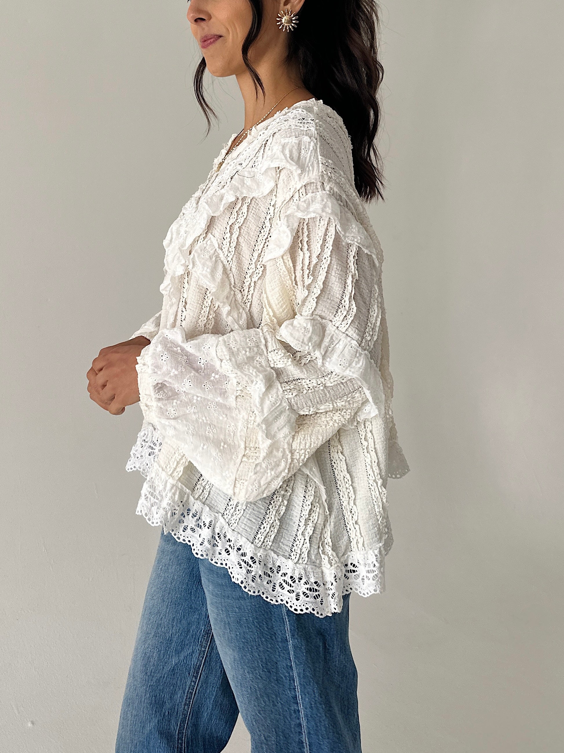 A woman wearing a white lace blouse with ruffles and blue jeans, shown from the side against a plain light gray wall.
