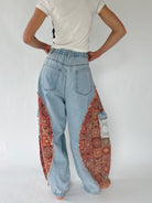 A person shown from the back wearing light blue wide-leg jeans with ornate red and gold patterned fabric panels integrated into the legs, paired with a white t-shirt against a plain white studio background.