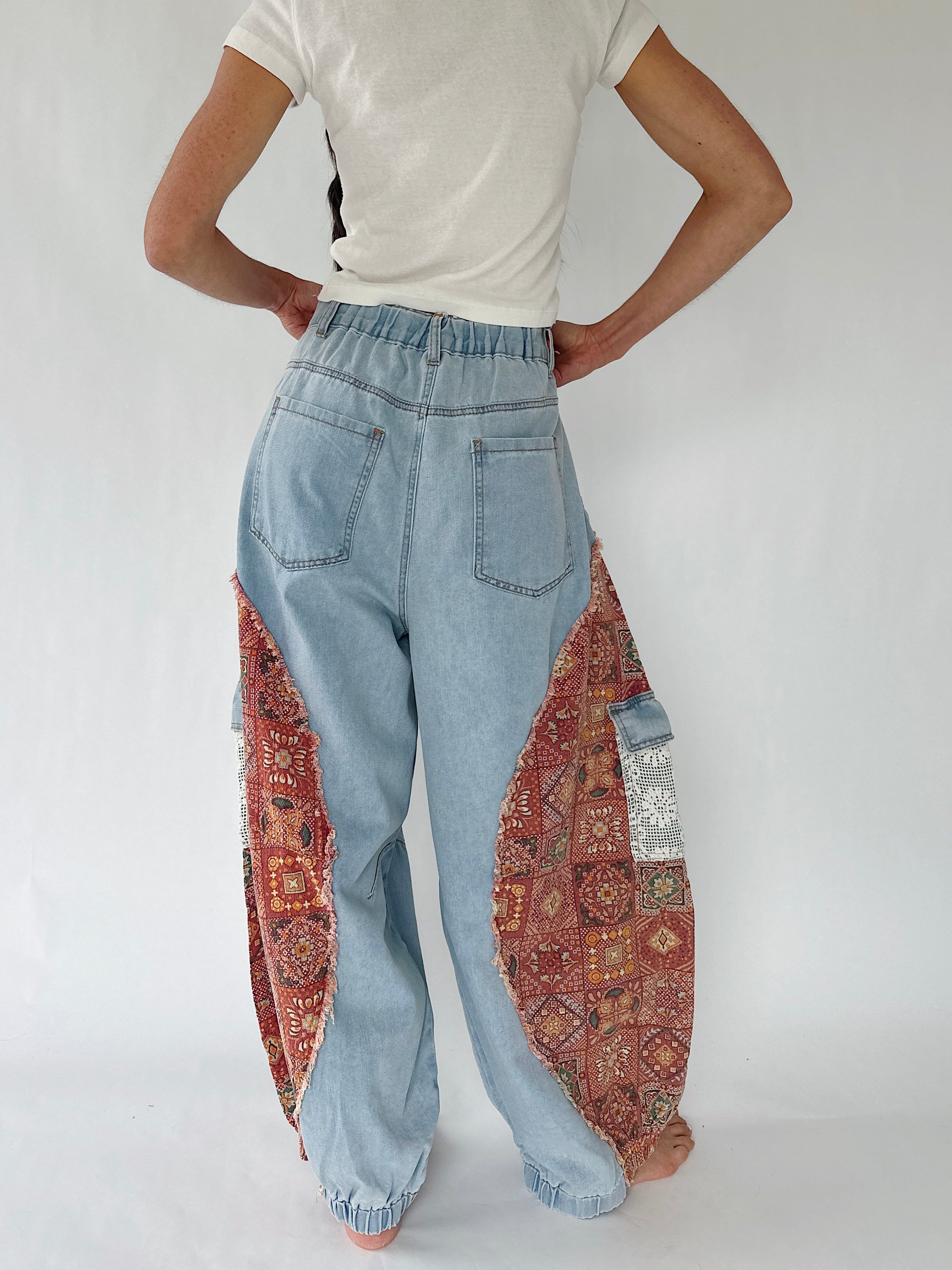 A person shown from the back wearing light blue wide-leg jeans with ornate red and gold patterned fabric panels integrated into the legs, paired with a white t-shirt against a plain white studio background.