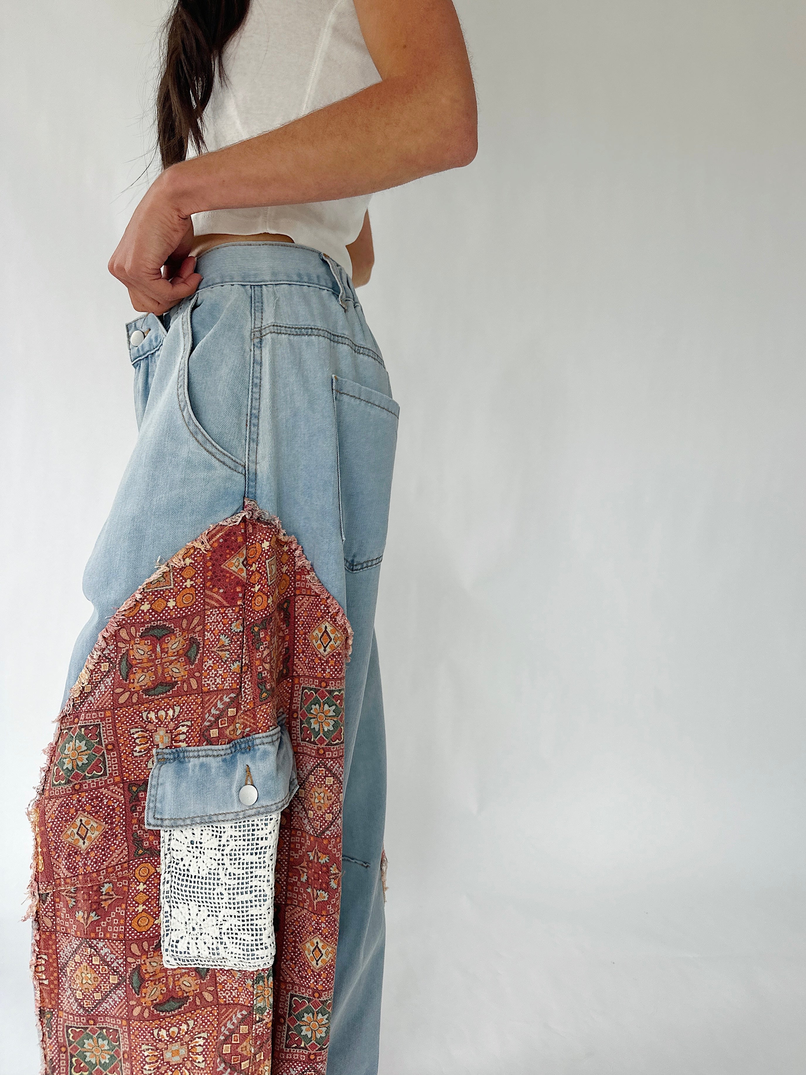 A person wearing light blue wide-leg denim jeans with a large rust-red patterned patchwork fabric panel and a small white sequined pocket detail, paired with a white cropped top against a plain light gray studio background.