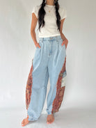 A model wearing light blue wide-leg denim pants with ornate red and gold patterned fabric panels on the outer sides, styled with a white t-shirt against a plain light background.