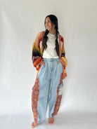 A woman stands barefoot against a plain light gray studio background, wearing a loose white top, light blue high-waisted jeans, and a long open-front cardigan with a multicolor gradient and paisley pattern.