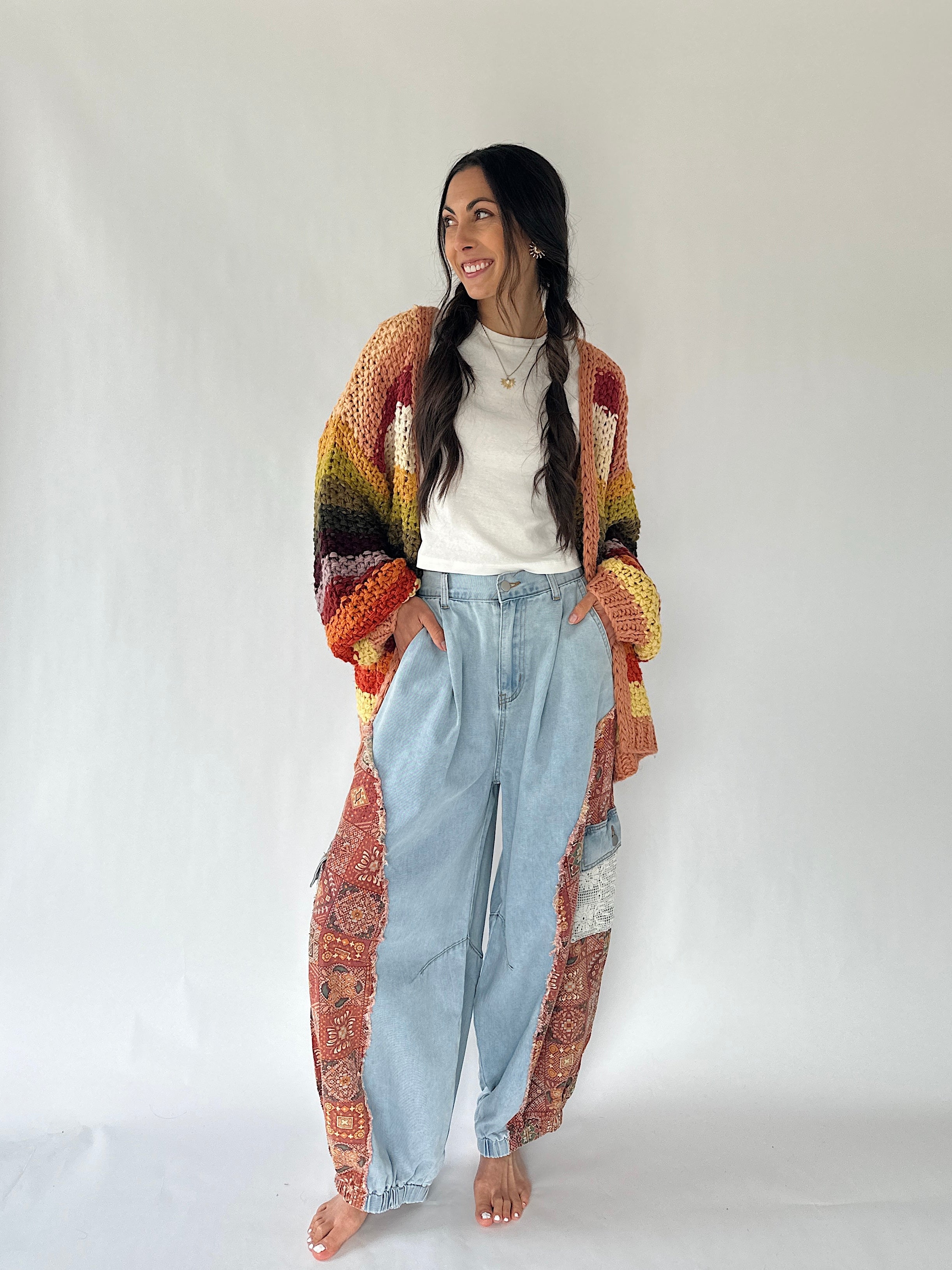 A woman stands barefoot against a plain light gray studio background, wearing a loose white top, light blue high-waisted jeans, and a long open-front cardigan with a multicolor gradient and paisley pattern.