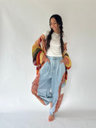 A woman modeling a colorful patchwork knit cardigan over a white t-shirt and light-wash wide-leg jeans, standing against a plain white studio background.