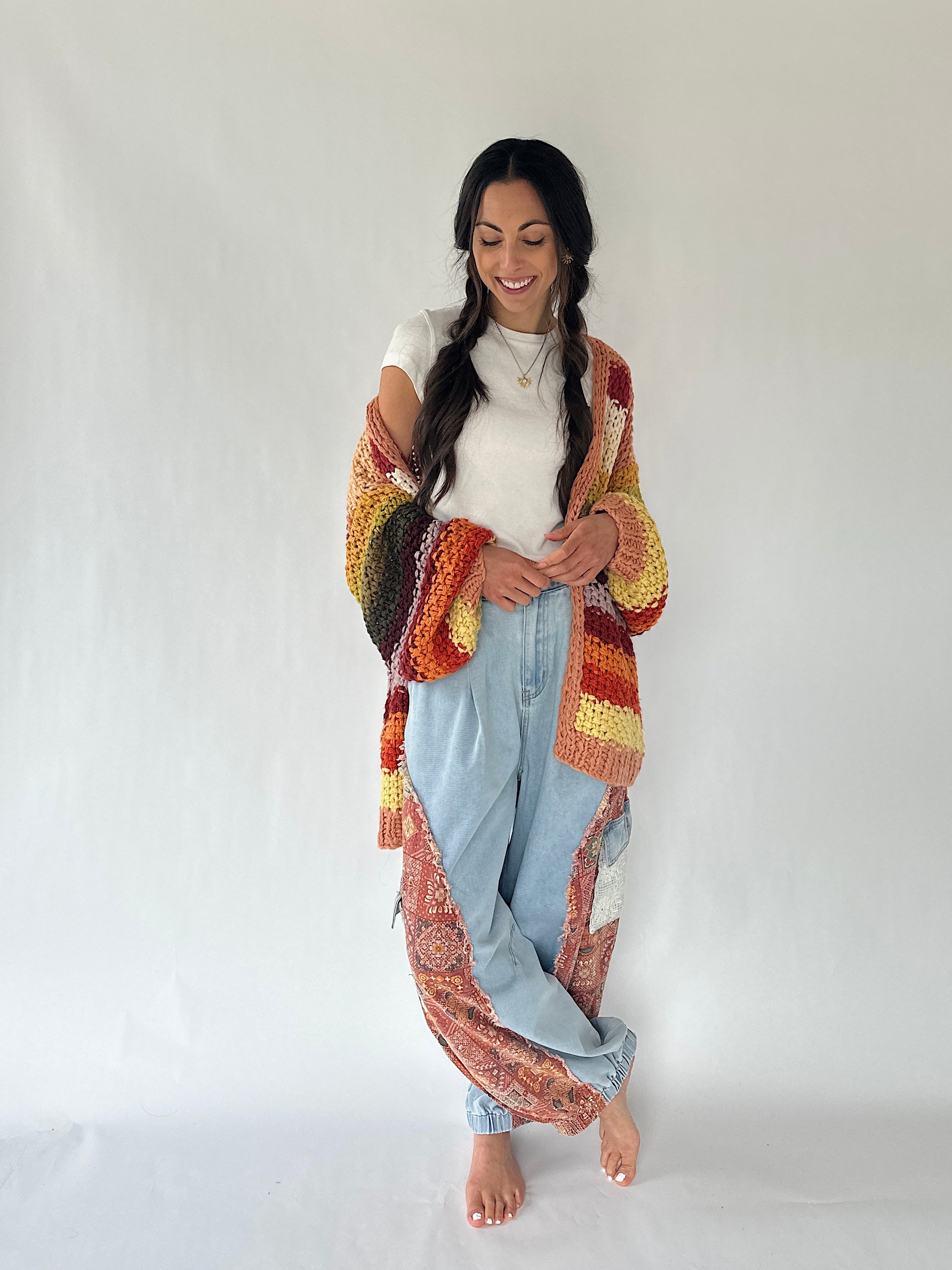 A woman stands barefoot against a plain white studio background, wearing a long, loose, patchwork-style cardigan or cover-up with a multicolor crochet and paisley pattern in orange, yellow, red, brown, and cream over a white top and light blue jeans.
