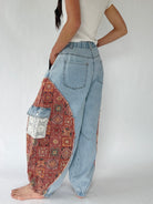 A person shown from the back wearing light-wash wide-leg jeans with large patchwork side pockets featuring rust-red patterned fabric and a small lace pouch pocket, against a plain light gray studio background.