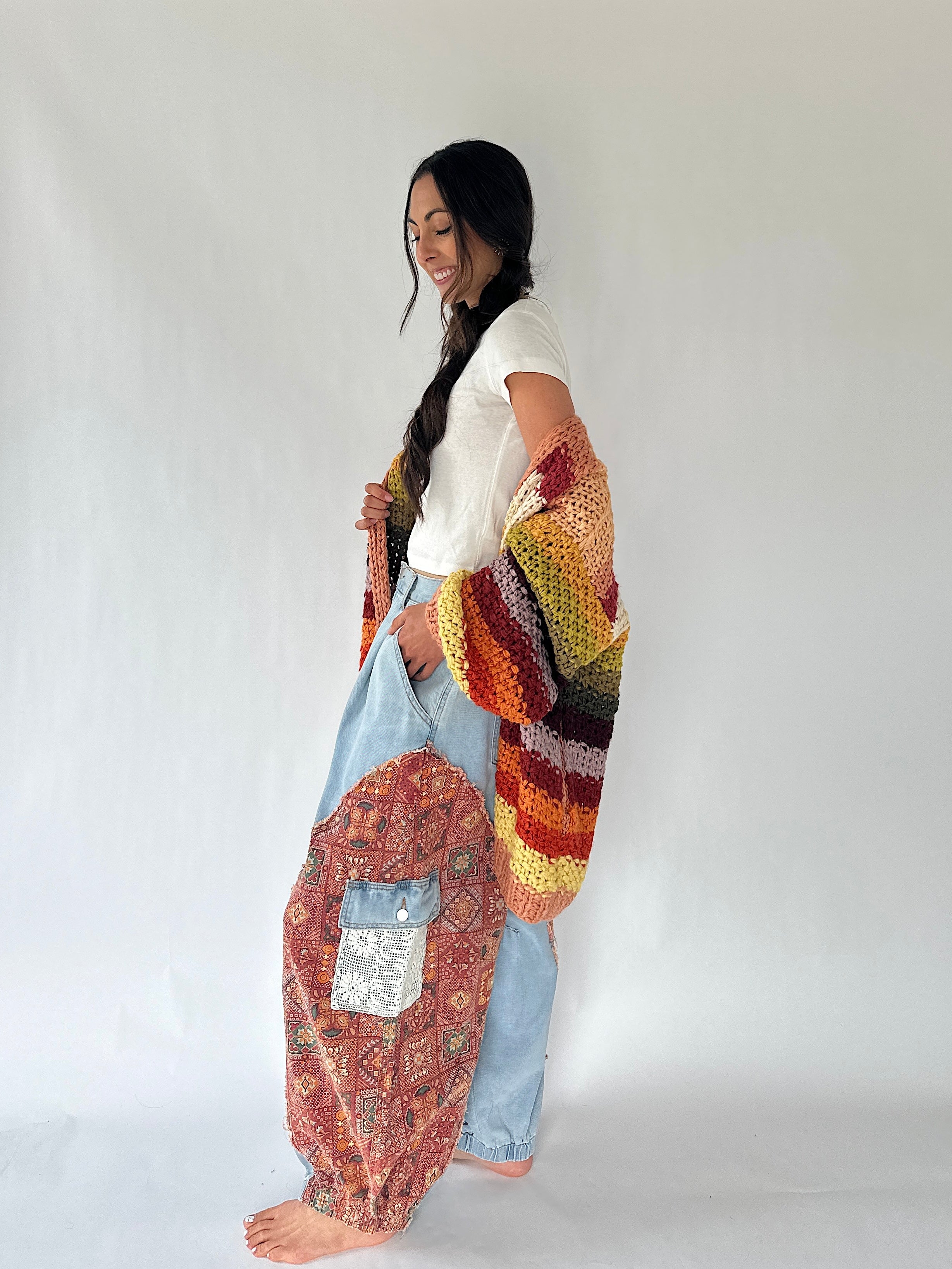 A woman stands barefoot against a plain light gray studio background, wearing a white short-sleeve top, light blue harem-style pants, and a large multicolored crocheted cardigan draped over her arm and around her body. The cardigan has a striped pattern in orange, yellow, green, brown, black, and cream.