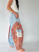 A person wearing light blue denim harem-style pants with large patchwork side panels in red, orange, and multicolor bohemian patterns, paired with a white sleeveless top, against a plain light gray studio background.