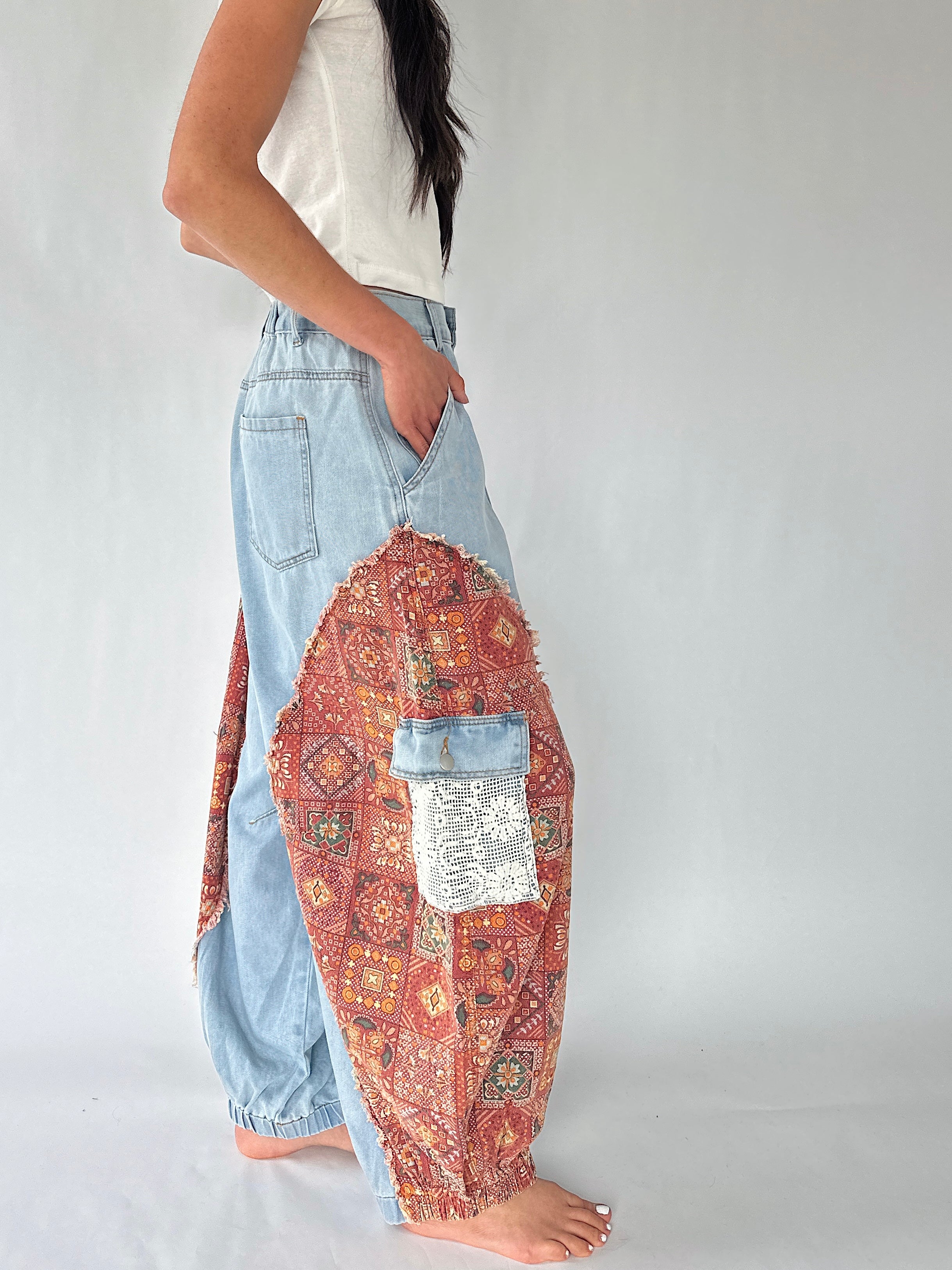 A person wearing light blue denim harem-style pants with large patchwork side panels in red, orange, and multicolor bohemian patterns, paired with a white sleeveless top, against a plain light gray studio background.