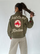 Person wearing a green jacket with 'West Coast Rodeo' text and cowboy logo on the back, standing against a white background.