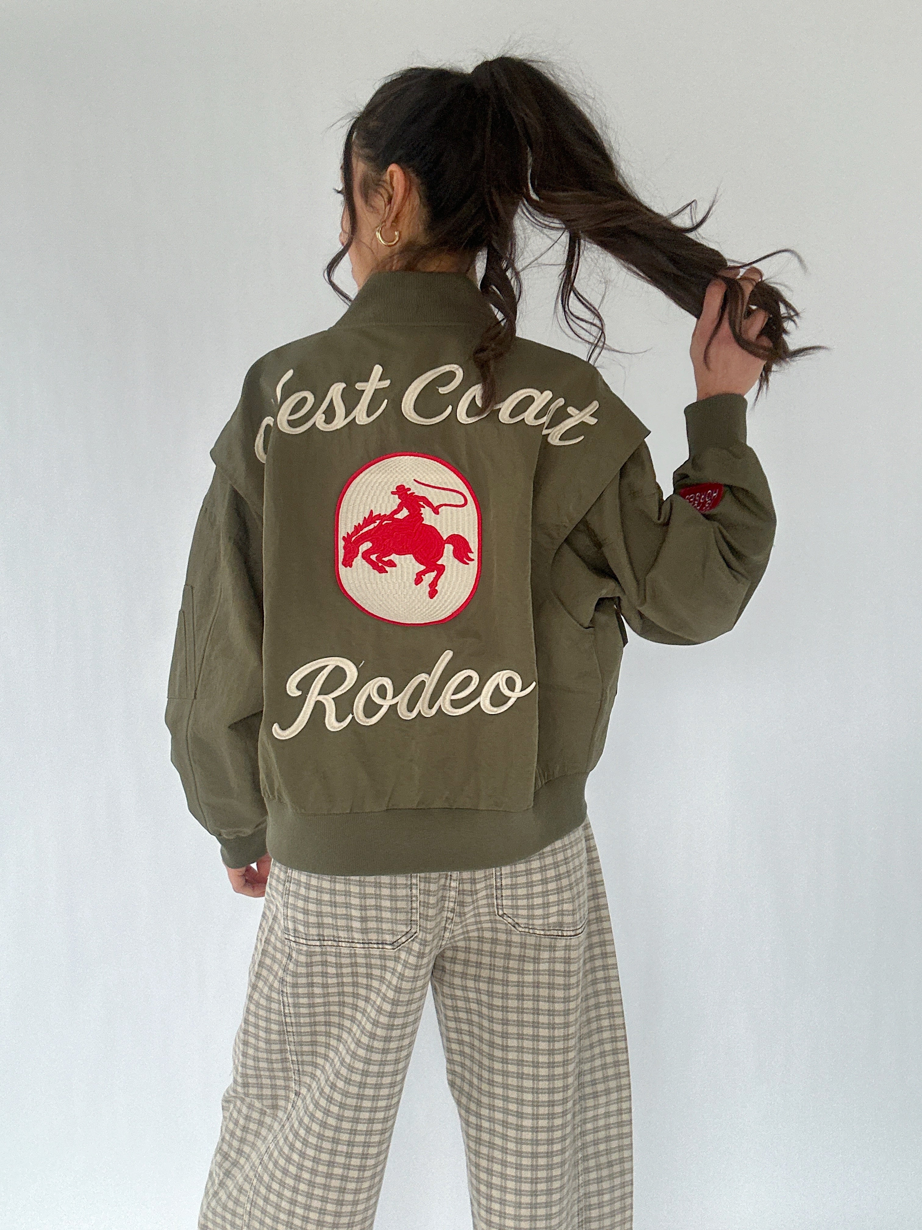 Person wearing a green jacket with 'West Coast Rodeo' text and cowboy logo on the back, standing against a white background.