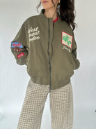 Person wearing a green bomber jacket with western patches and text, standing against a white background